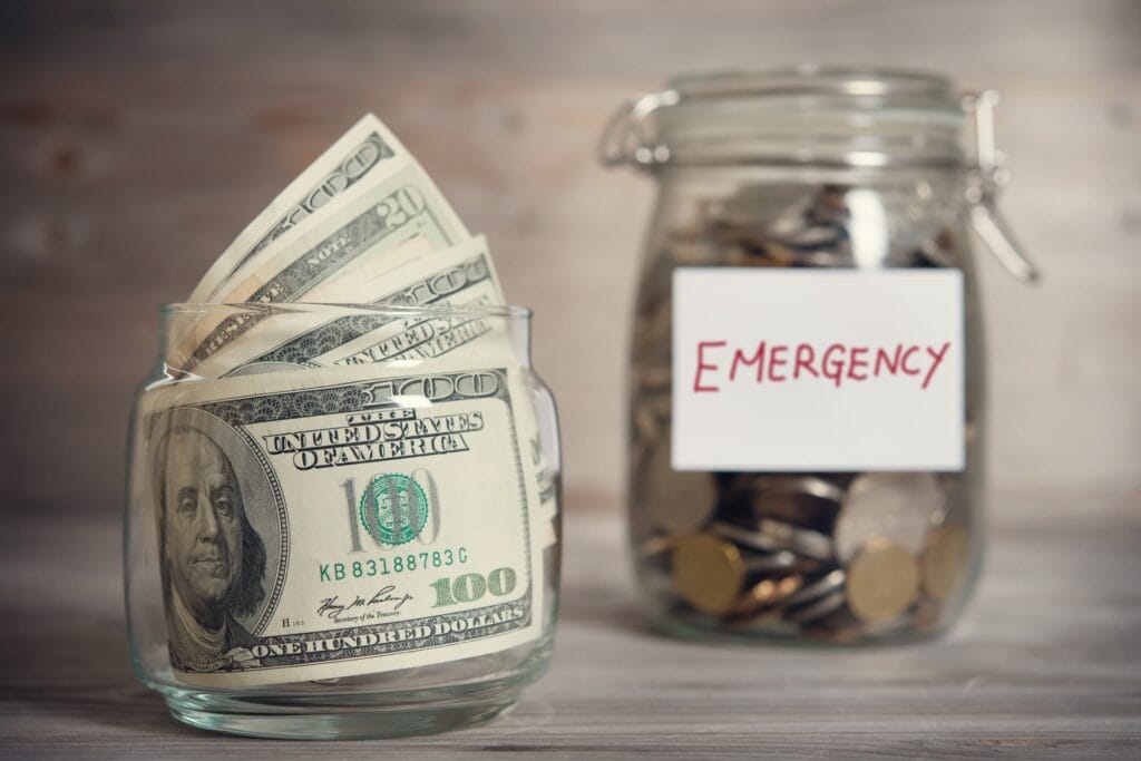 A stack of $100 bills and a jar filled with coins with "Emergency" written on the front.