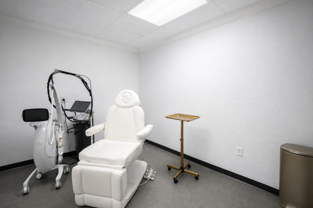 An empty med spa treatment room with a white reclining chair and a brass rolling tray
